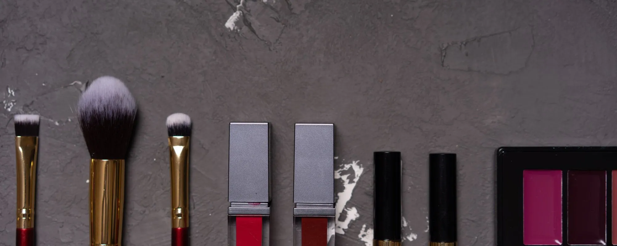 Kuas, lipstik, dan palet makeup berjajar di atas latar gelap