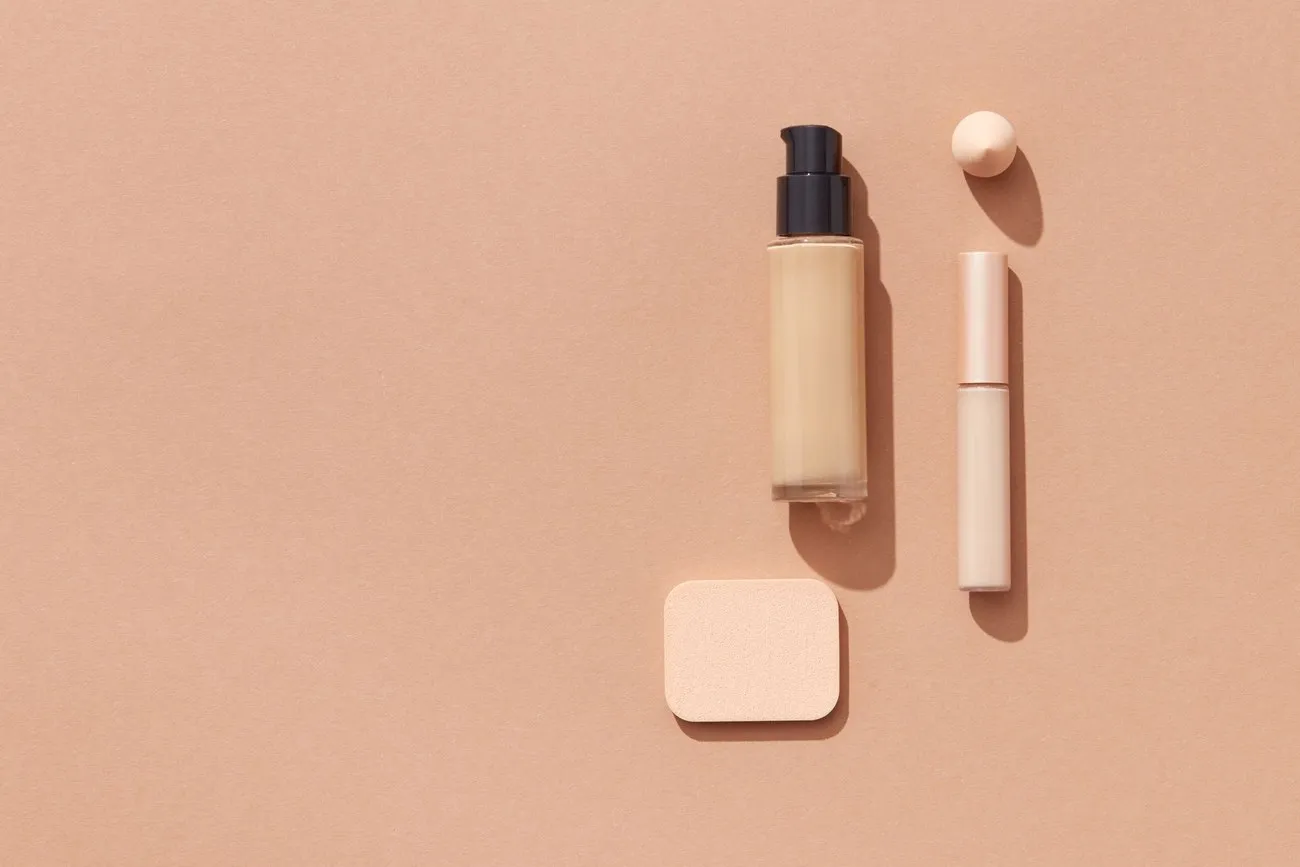 Botol alas bedak, concealer, dan spons di atas latar jingga muda
